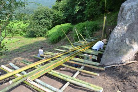 傾斜地に配置された複数の竹が足場のように並べられ、奥にいる二人の作業者が取り付け作業をしている様子の写真