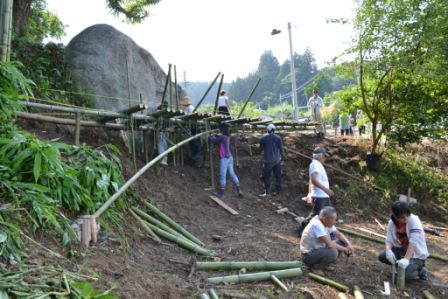 竹の骨組みが立ち上がり始め、斜面で何人もの人々がそれぞれの持ち場で作業を進めている様子を写した写真