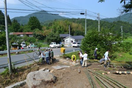 道路のそばで複数の作業グループが竹を切ったり並べたりして作業している様子を写した写真