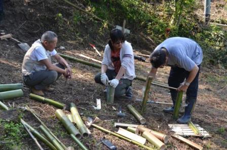 地面に座り込んで竹をのこぎりで切ったり、寸法を合わせて加工している3人の男性が、真剣な表情で作業している様子の写真