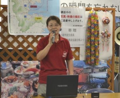 赤いポロシャツを着た菅原清香さんがマイクをもって話をしている様子の写真