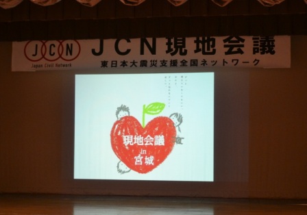 ステージ上のスクリーンに、赤いハートのイラストと「現地会議 in 宮城」の文字が大きく映されており、その上には「JCN現地会議 東日本大震災支援全国ネットワーク」の横断幕が掲げられているJCN現地会議のステージ正面の写真