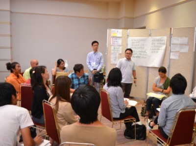 小グループのセッションで、参加者たちが椅子を囲んで座り、模造紙にまとめられた意見を見ながら、立っている男性の話に耳を傾けている様子の写真