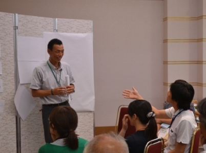 前に立つ男性が笑顔で話しながら、向かい側の若者が手を挙げて意見を述べており、活発な対話が行われている様子の写真