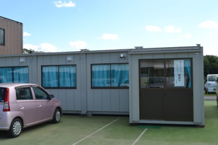 青いカーテンのある窓が並ぶグレーのプレハブ建物で、、「ネイチャーセンター準備室」と書かれた紙が玄関扉に貼られている準備室の外観の写真