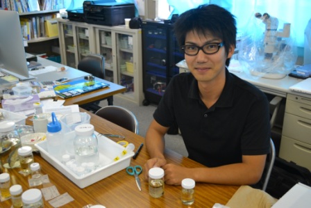 アルコール標本を前にして机に向かって座り、にこやかにこちらを見ている黒いシャツの黒田和真さんが写っている準備室での作業の写真