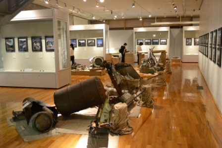 錆びたドラム缶のようなものや金属製の破片が床に並べられ、壁には多数の写真が展示されている展示室の全景の写真