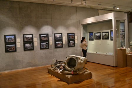 壁に掛けられた震災と津波の被害を撮影した写真パネルと、展示物の大きな金属破片のそばで展示物を見ている来館者が写っている展示室の写真