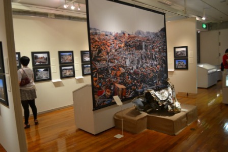 震災で崩壊した街並みの巨大な写真パネルと瓦礫の展示が中央にあり、周囲の壁には震災後の風景写真が額装されて展示されている室内展示室の写真