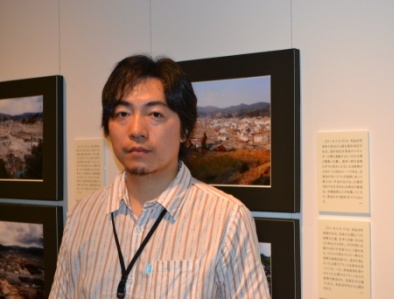 展示写真の前に立つ学芸員の岡野志龍さんの写真