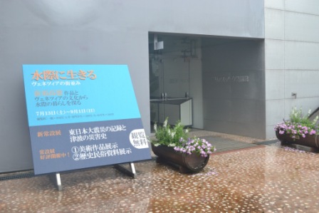 「水際に生きる」と題された青い展示案内板が雨に濡れた地面に立てられ、その奥に展示施設の入口が見える屋外の様子の写真