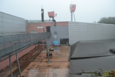 霧に包まれた展示施設の外観を高所から見下ろし、赤い監視塔や連絡通路、濡れた中庭と展示入口が確認できる建物全体の様子の写真