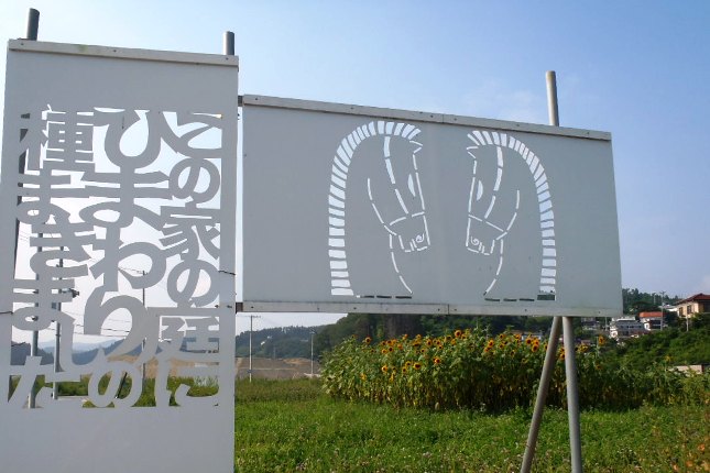 「『この家の庭にひまわりの種をまきました』という文字が抜かれたきりこの白い看板と、馬の絵が描かれたきりこの看板の後ろに、ひまわり畑が広がる様子の写真