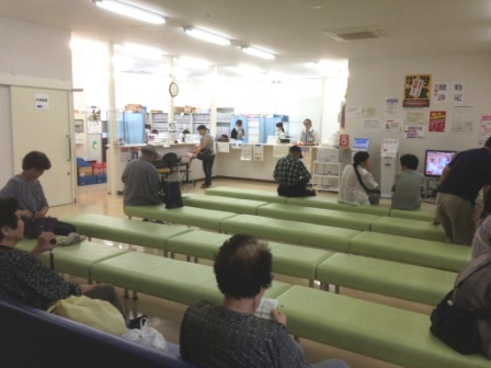 緑色の長椅子が規則的に並べられた待合室で、高齢者を中心とした複数の患者が座って診察の順番を待ち、奥のカウンターでは職員が受付対応をしている診療所内の待合室の写真