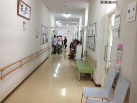 白い壁と明るい照明の廊下の両側に診療科の出入口が並び、壁には手すりが設置され、高齢の患者たちが長椅子に座って順番を待っている診療所内の廊下の写真