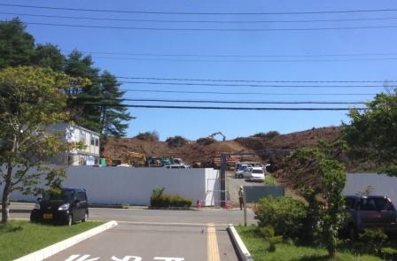 奥の丘の上で重機や作業車両が動いており、白い仮囲いとプレハブが設置されている高台の工事現場を写した写真