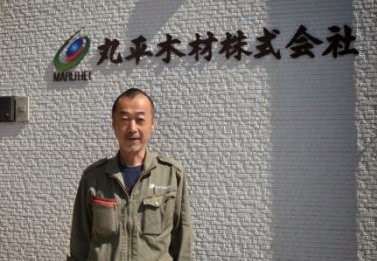 白い外壁に『丸平木材株式会社』の社名看板が掲げられた建物の前に立っている、作業服を着た小野寺邦夫社長の写真