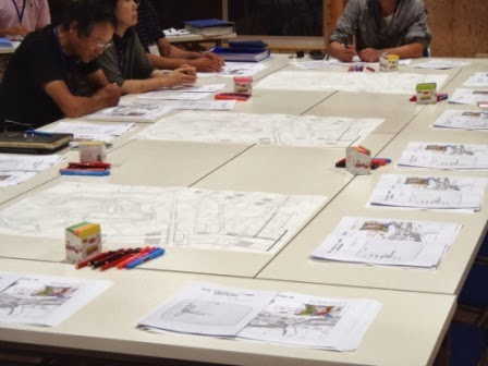 会議用の白いテーブルの上に、地域の地図や図面とともに、赤青のペンや色鉛筆、資料が並べられ、話し合いが行われる机の上を写した写真