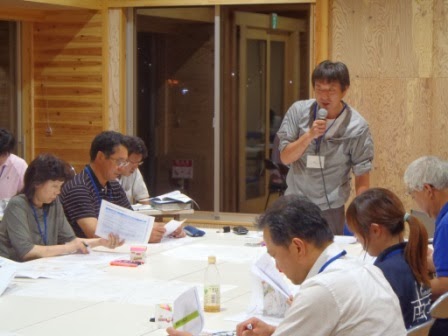 中央の男性が立ち上がってマイクで発言しており、周囲の参加者が資料を見ながら耳を傾けている会議の様子の写真