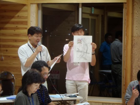 眼鏡をかけた男性がマイクを持って資料を読み上げ、隣の若い男性が地図を全員に見えるよう高く掲げている会議の様子の写真