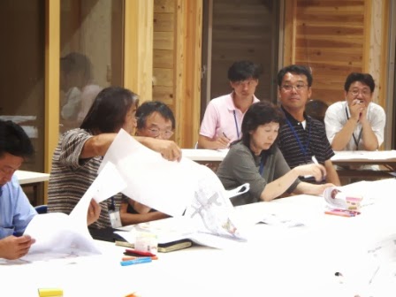 参加者たちが白いテーブルを囲んで座り、一人の参加者が地図を広げて隣の男性に見せながら話し合い、他の人たちは真剣な表情で資料を見つめている様子の写真