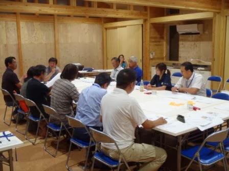 机のまわりで参加者が着席し、資料を手にしながら静かに話を聞いている様子の写真