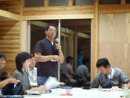 眼鏡をかけた男性が立ち上がってマイクで意見を述べ、周囲の人たちがうなずいたり資料を見たりして耳を傾けている、積極的な意見交換の写真