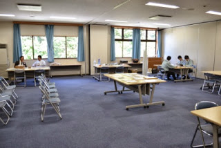 窓際に青いカーテンがかかる広い会議室で、数組の住民と職員が向かい合う形で長机に着いて相談しており、その手前には相談の順番を待つための椅子が整然と並んでいる相談会場の全体の様子の写真