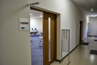 廊下に面した大きな会議室の入口ドアが開いており、受付案内の立て看板が設置され、その奥で相談対応が行われている相談会場の外観の写真