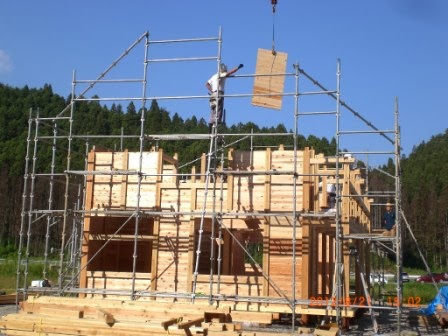 木材で組み上げられた建物の骨組みの中で、作業員が足場を使ってクレーンから吊るされた木材を取り付けようとしており、木造建築の建設が進む様子がわかる写真