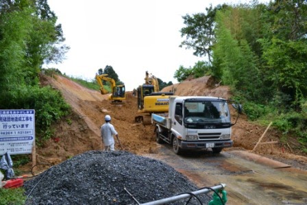 周囲を木々に囲まれた斜面状の地形を2台の黄色いショベルカーが掘削作業を行い白いトラックが停車し、そのそばにヘルメットを着けた作業員が立っている写真