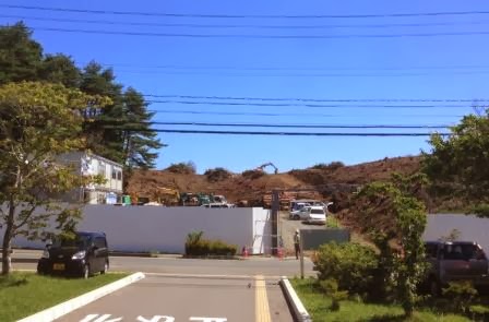 現場全体は白い塀で囲まれており、塀の奥には複数の車両が停まっており乗用車と工事用の重機が混在し背景には緩やかな傾斜のある小高い丘があり、黄色いショベルカーが作業をしている様子の写真