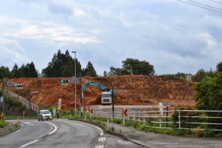 道路の側の小高い丘のような地形の中で重機が土を掘り稼働しており、工事が進行中である様子を示している工事中の写真