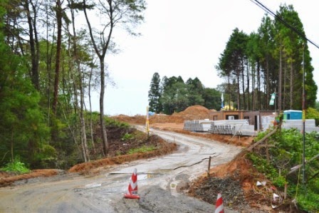 豊かな森林に囲まれた静かなぬかるんだ地面の山道が中央に伸びており、道の両側には木々が密集して立ち並び、右側には現場事務所や工事の材料が置かれた写真