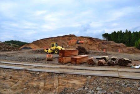 山のように盛り上がった小高い場所でショベルカーが土を掘削しており、その下の平地ではブルドーザーが整地作業を行い、周囲には木材などの資材が置かれている工事現場の写真