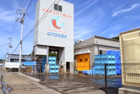 「よみがえれ！南三陸」と赤字で書かれた建物にサーモンのイラストと「GYOUBA」の文字が描かれ、手前にはたくさんの青いコンテナが積まれている写真