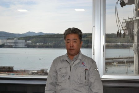 外に海と港の風景が広がる窓の前に立つ、作業服を着た高橋正宜社長の写真