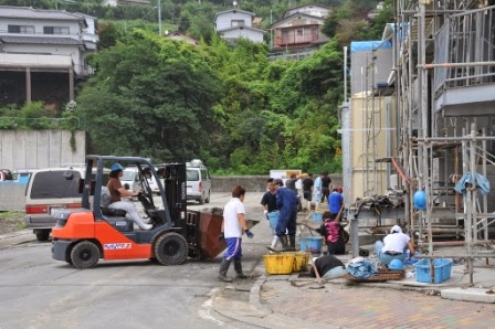 足場の組まれた建物の前では複数の人が作業を行い、傍でオレンジ色のフォークリフトが荷物を運んでいる様子の写真