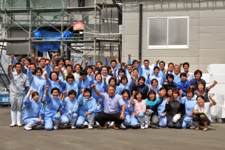 足場が組まれた建物の前に社員の皆さんが集まり、笑顔でガッツポーズをして写っている集合写真