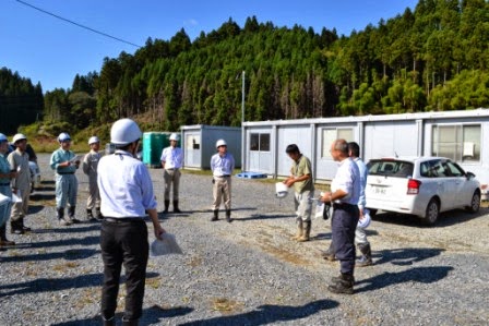 プレハブの建物の前で工事関係者らが円になって打ち合わせを行っている写真