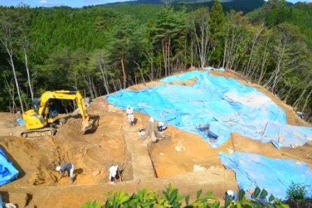 重機と作業員が地面を掘り進めている傍らで、広い範囲に青いビニールシートが掛けられている山中の発掘現場の全景写真