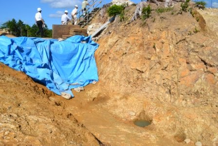 削られた岩肌の下に青いビニールシートがかけられ、上部に数人のヘルメットをかぶった作業員が立っている発掘調査現場の様子の写真