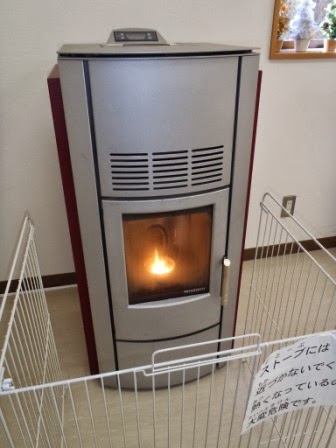 銀色のペレットストーブが室内に設置され、正面のガラス越しに炎が見えていて、周囲には安全のための白い柵が設置されている写真