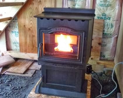 壁に断熱材が施工された建築中の室内で黒いペレットストーブが稼働しており、前面のガラス越しに赤く燃える炎が見える写真