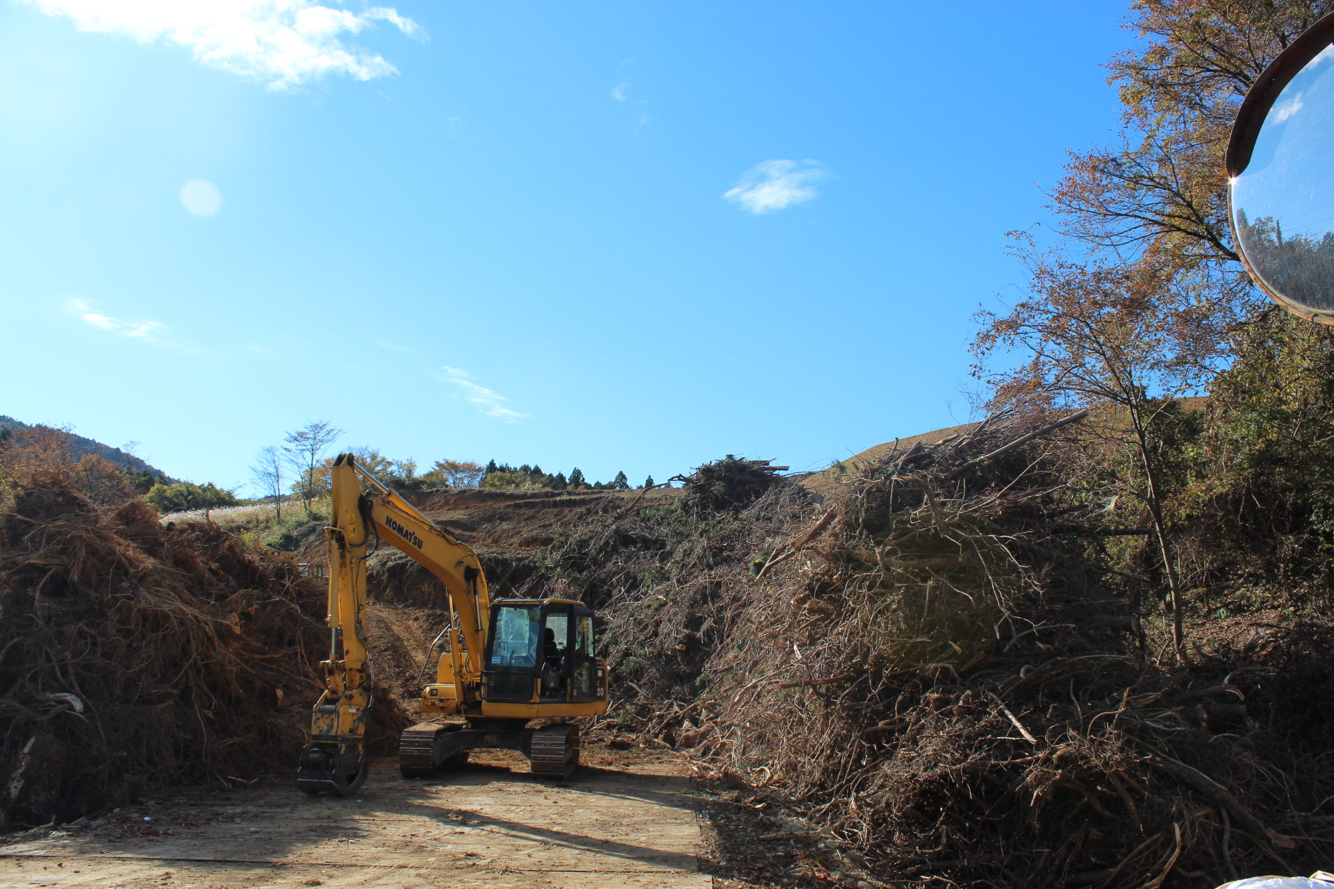 山間の造成地で重機が伐採された木々の山を前に作業している様子を写した写真