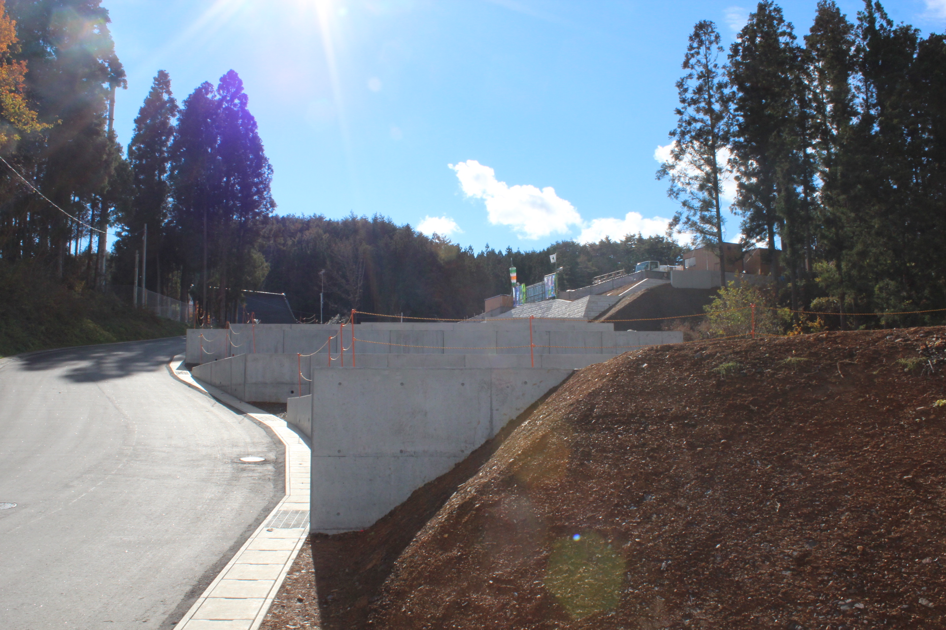 快晴の空の下、傾斜地に沿ってコンクリートの擁壁と舗装道路が整備されている工事現場の写真