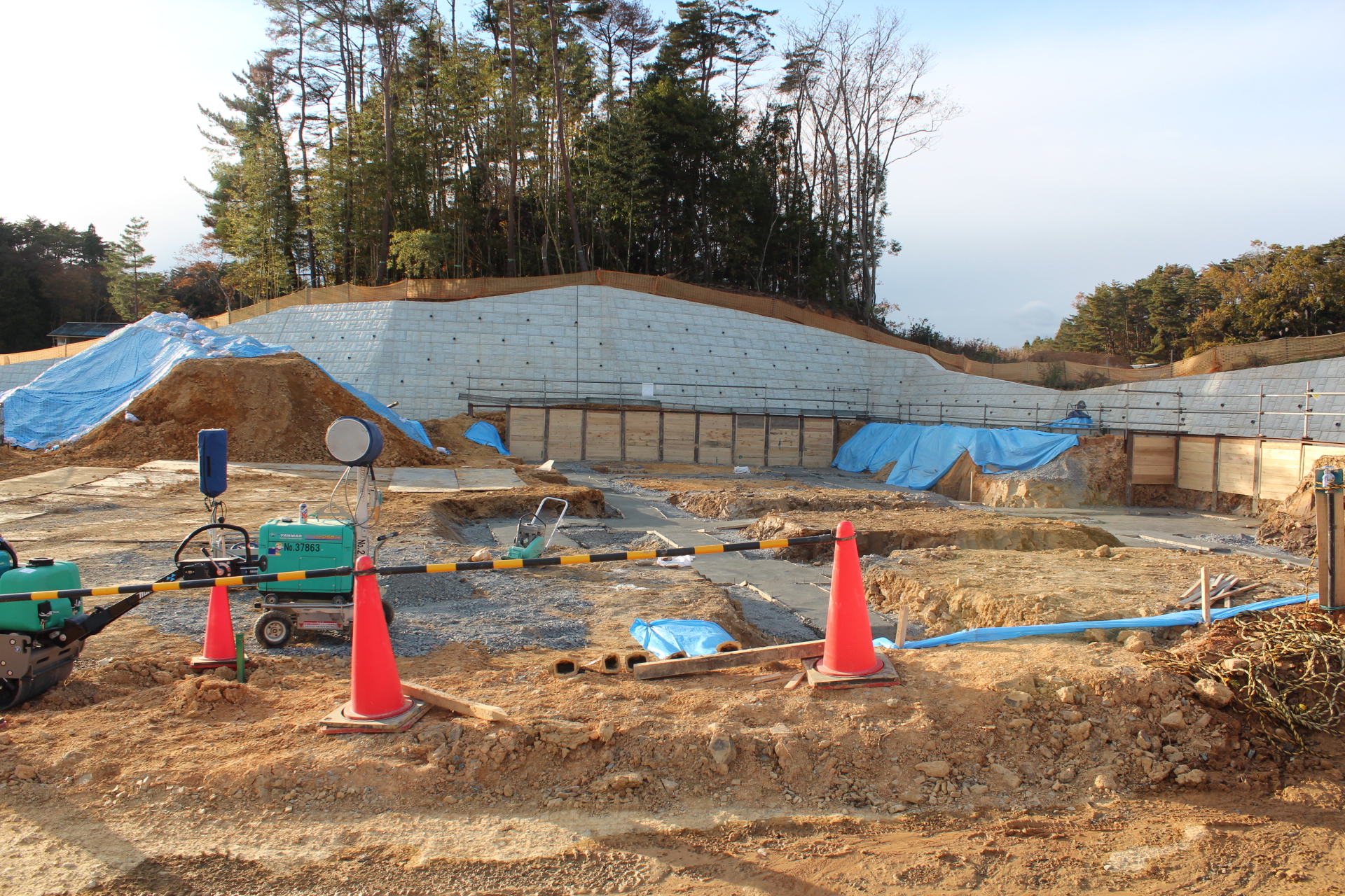 斜面に造成された広い敷地で地盤の整備とブルーシートによる覆いが施され、背後に竹林と擁壁が見える造成工事中の写真