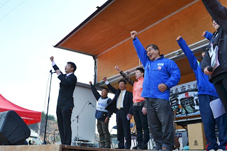 仮設ステージの上で複数の関係者が拳を掲げ、開催スタートの合図をしている様子の写真