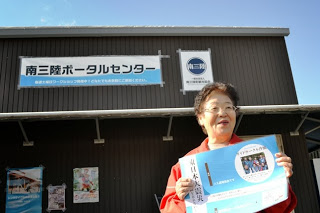 「南三陸ポータルセンター」の建物前で、赤いジャンパーを着た女性が笑顔でチラシを掲げている様子を写した写真