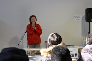 赤いジャンパーを着た女性がマイクを持って壇上で話し、来場者たちが椅子に座って耳を傾けている様子を写した写真